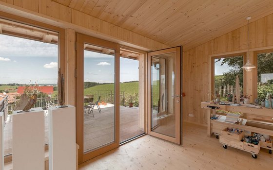 Helles, freundliches Atelier in einem modernen Holzhaus mit großen Fenstertüren, die auf einen Balkon mit Ausblick auf grüne Felder führen. Im Raum sind eine Arbeitstischreihe und verschiedene Kunstskulpturen zu sehen.