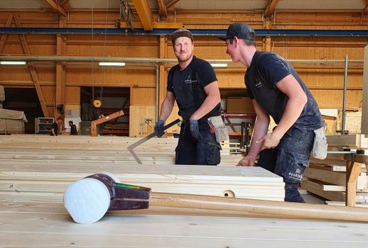 Zwei Mitarbeiter von Rehklau arbeiten in einer großen Werkstatt an Holzplatten. Sie tragen Arbeitskleidung und wirken konzentriert bei der Arbeit mit einem Winkelmaß. Im Vordergrund liegt ein großer Hammer auf dem Tisch.