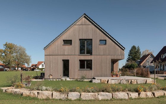 Modernes, minimalistisches Holzhaus in einem Wohngebiet, umgeben von einem naturbelassenen Steingarten mit blühenden Pflanzen.