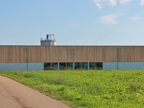 Ein langgestrecktes Gebäude mit einer Holzfassade aus Rehklau Vollholz, das auf einer großen Wiese steht. Im Hintergrund ist ein Silo sichtbar, das die industrielle Nutzung des Gebäudes andeutet. Dieses Bild zeigt eine der Referenzen von Rehklau Vollholz.