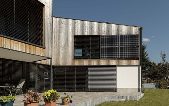 Ein modernes Holzhaus mit grauem Giebeldach und Solarpaneelen auf der Dachfläche. Das Haus steht in einem grünen Garten mit einer möblierten Terrasse und umgeben von Bäumen.