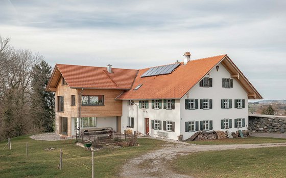 Ein großes Landhaus mit einer Kombination aus weißem Putz und Holzfassade, rotem Ziegeldach und grünen Fensterläden. Auf dem Dach befinden sich Solarpaneele, und der umgebende Hof ist ländlich und weitläufig.