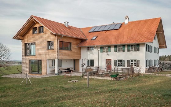 Eine moderne Renovierung eines Bauernhauses mit einer Erweiterung aus Holz auf der linken Seite und einem traditionellen weißen Gebäude mit grün lackierten Fensterläden auf der rechten Seite. Das Gebäude hat ein rotes Ziegeldach, und im Vordergrund liegt eine weitläufige Wiese.