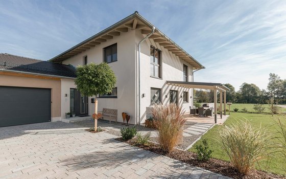 Ein zweistöckiges modernes Haus mit hellgrauer Fassade und angebauter Garage, umgeben von einem gepflasterten Vorplatz. Vor dem Eingang steht ein Baum, und eine seitliche Terrasse mit Pergola bietet Sitzgelegenheiten im Garten