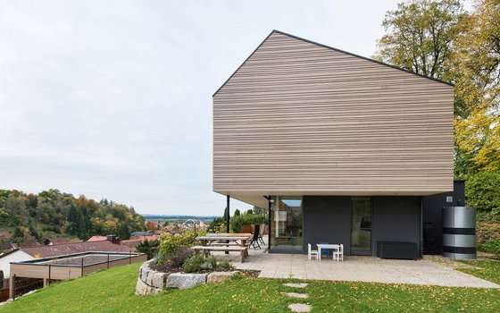 Wohnhauses mit auffälliger, überhängender Oberstruktur, vertikaler Holzfassade und großflächigen Glasfronten. Vor dem Haus befindet sich eine Terrasse mit einem Holztisch. Das Gebäude steht auf einer Anhöhe, die einen malerischen Ausblick auf das umliegende Dorf und die herbstliche Landschaft bietet.