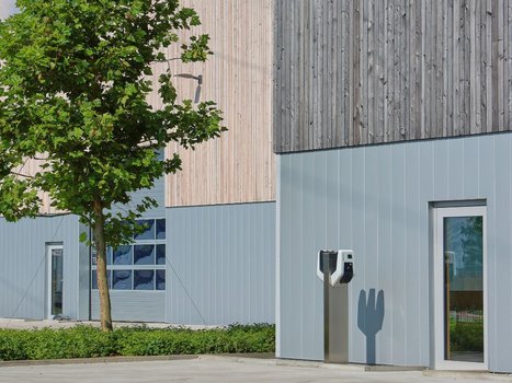 Nahaufnahme einer modernen, neu gebauten Produktions-/Lagerhalle mit einer E-Ladestation im Vordergrund. Ein Baum spendet Schatten, was die umweltfreundliche Ausrichtung dieser Rehklau Vollholz Referenz unterstreicht.