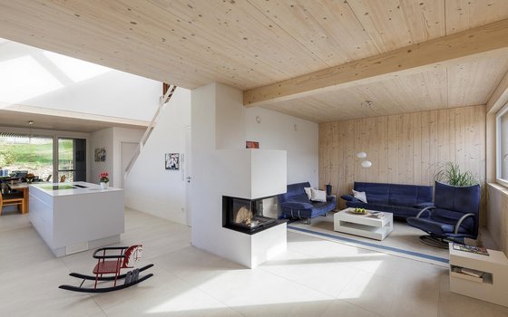 Helles und luftiges Wohnzimmer mit natürlicher Holzverkleidung und hohen Decken. Die Einrichtung umfasst bequeme blaue Sofas, einen modernen Kamin als Raumteiler und eine minimalistische weiße Kücheninsel