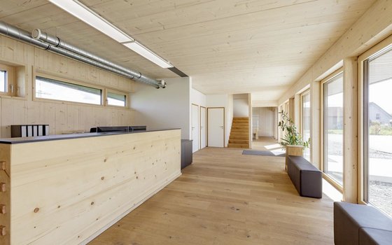 Ein heller Empfangsbereich mit Holzelementen, großen Fenstern und einem minimalistischen Design. Eine Treppe führt in das obere Stockwerk.
