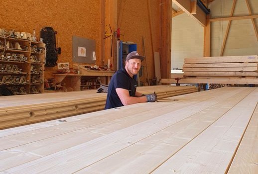 Ein lachender Mitarbeiter in der Rehklau-Werkstatt, umgeben von großen Holzbrettern. Der Raum vermittelt eine warme Atmosphäre durch die Verwendung von Holzmaterialien.