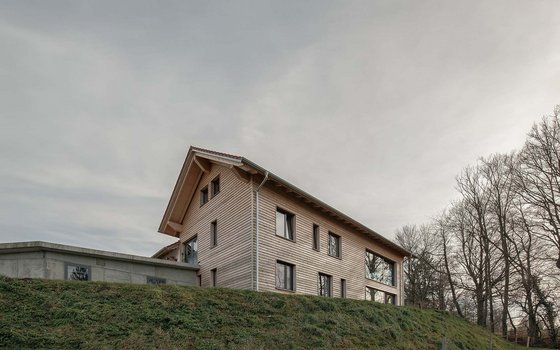Ein modernes Holzhaus mit Walmdach, das auf einer kleinen Anhöhe steht. Die schlichte Fassade ist mit Holz verkleidet, und das Gebäude ist von einer Wiese und hohen Bäumen umgeben.