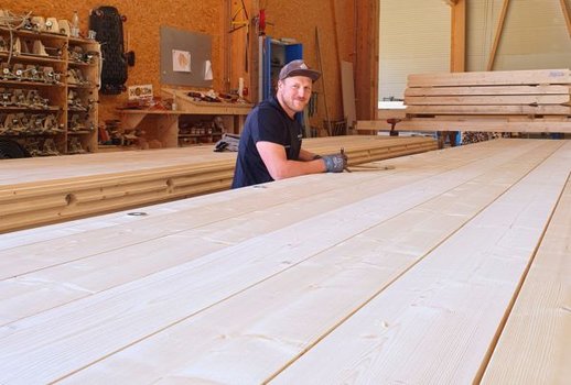 Ein Mitarbeiter in der Rehklau Werkstatt steht an einem großen Holzbrett und lächelt in die Kamera. Der Raum ist mit Werkzeugen und Holzbalken ausgestattet.