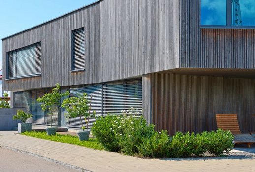 Außenansicht eines zweistöckigen Gebäudes von Rehklau mit Holzfassade und Pflanzen im Vorgarten. Das Design wirkt minimalistisch und naturverbunden.