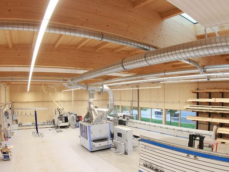 Eine geräumige, helle Werkstatt mit einer Decke und Wänden aus Rehklau Vollholz. Große Maschinen und Belüftungssysteme sind zu sehen, die diese Rehklau Vollholz Referenz in einer modernen Arbeitsumgebung verorten.