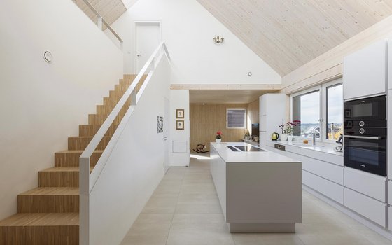 Moderne Küche in einem Haus mit offener Raumaufteilung, ausgestattet mit einer weißen Einbauküche, Zentralinsel und Holztreppen, die zu einer oberen Ebene führen, unter einer Decke mit sichtbaren Holzbalken.
