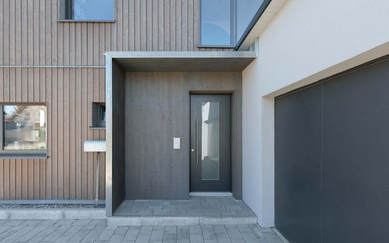 Modern gestalteter Hauseingang mit grauer und holzerner Fassade, minimalistischer Tür und schlichtem Betonvordach, der die Attraktivität des Eingangsbereichs steigert.