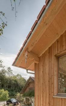 Detailaufnahme der Holzverkleidung und Dachkonstruktion eines modernen Wohnhauses, mit traditionellen Verzierungen und Kupferdachrinnen. Der Hintergrund zeigt einen älteren, ebenfalls charmanten Gebäudeteil und Bäume, die den ländlichen Charakter unterstreichen.