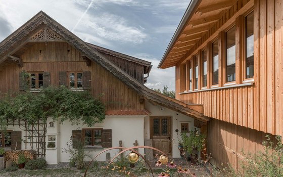 Ein traditionelles Bauernhaus mit einer Holzkonstruktion und einem hellen Putz. Der rechte Teil des Hauses ist aus Holz und verfügt über große Fenster, während der linke Teil im klassischen alpenländischen Stil gestaltet ist. Im Vordergrund befindet sich ein dekorativer Garten mit einem Metallkunstwerk und einigen Pflanzen.