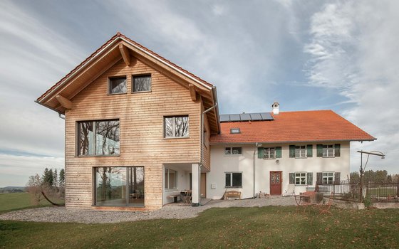 Ein ländliches Anwesen mit einer Kombination aus moderner und traditioneller Architektur, darunter ein neuer Anbau mit Holzverkleidung und großflächigen Fenstern neben einem klassischen weißen Gebäude mit grünen Fensterläden und einem roten Ziegeldach, umgeben von einer weiten Landschaft unter bewölktem Himmel.