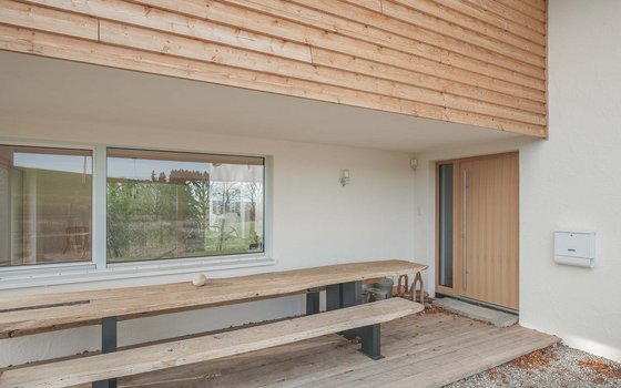 Ein schlicht gestalteter Eingangsbereich eines modernen Hauses mit einer rustikalen Holzbank unter einer überdachten Terrasse.