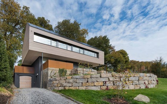 Modernes, architektonisch gestaltetes Haus in Schwebekonstruktion über einem Garagengrund mit großen Panoramafenstern und einer umgebenden Steinterrasse.