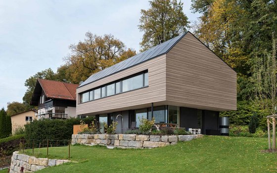 Moderner Anbau an ein traditionelles Haus, gelegen in einem üppig grünen Garten. Der Anbau zeigt eine minimalistische Architektur mit einer großen Fensterfront und Holzverkleidung, perfekt integriert in eine natürliche Umgebung.