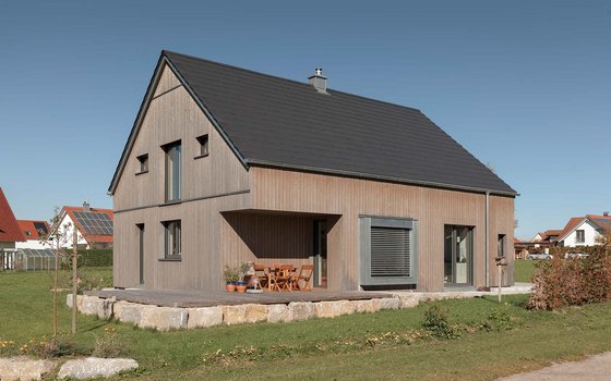 Modernes Einfamilienhaus mit grauer Holzverkleidung und einer großen überdachten Terrasse, umgeben von einem naturnahen Garten.