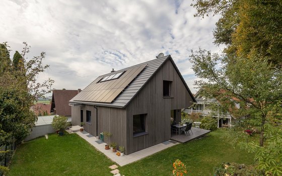 Ein modernes Holzhaus mit großen Glasfenstern und einer kleinen Kiesfläche um das Gebäude. Das Haus bietet einen Blick auf eine ländliche Umgebung mit Feldern und Bäumen.
