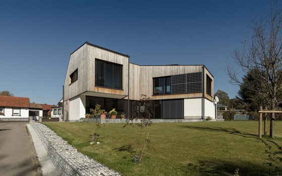 Kubusartiges Haus mit moderner Holzverkleidung und großen, getönten Fenstern. Die schlichte, kantige Architektur wird durch den großzügigen Garten ergänzt.