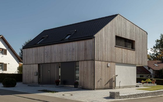 Modernes zweistöckiges Haus mit vertikaler Holzverkleidung, Schieferdach und großen Fenstern, gelegen in einer ruhigen Wohngegend.