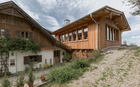 Bauernhaus mit modernem Anbau aus Holz, gezeigt in einer ländlichen Umgebung. Der alte Teil des Hauses zeichnet sich durch traditionelle Holzschindeln und verschachtelte Fenster aus, während der Anbau große Fenster und eine klare Holzfassade bietet.