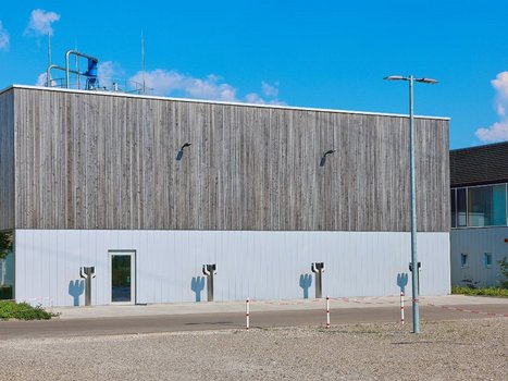 Ein modernes Gebäude mit einer hohen Holzfassade unter blauem Himmel. Das Gebäude ist umgeben von Kies und einem Pflasterstreifen und repräsentiert eine der Rehklau Vollholz Referenzen.