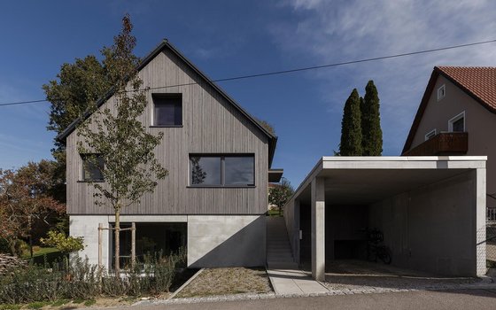 Ein modernes Einfamilienhaus mit Holzfassade und Carport aus Beton, in einem ruhigen Wohngebiet. Das Haus hat ein spitzes Dach und große Fenster, die einen offenen Blick auf den Vorgarten und die Umgebung bieten.