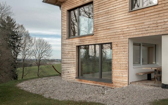 Detailansicht eines Holzhauses mit vertikaler Holzverkleidung und großen Glasfronten, die einen weiten Blick auf die umliegende Landschaft bieten. Der Bereich um das Haus ist mit Kies bedeckt.