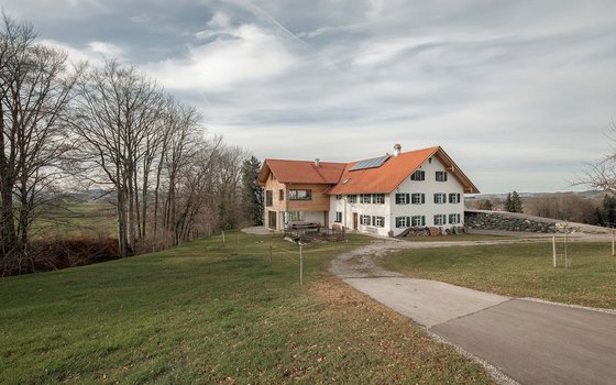 Ein traditionelles Bauernhaus mit rotem Ziegeldach und grünen Fensterläden, umgeben von weitläufiger Landschaft. Vor dem Haus befindet sich eine Rasenfläche und einige Bäume.