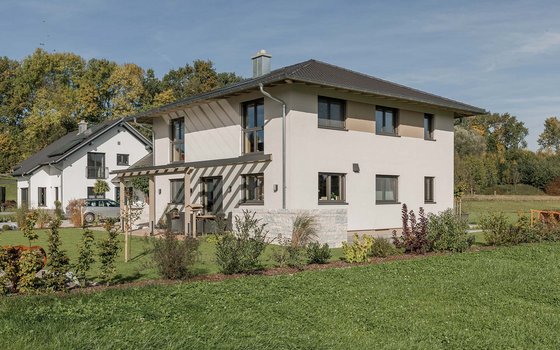 Ein zweistöckiges modernes Haus mit weißer Fassade, großen Fenstern und einem gepflegten Garten. Ein angrenzender Sitzbereich im Freien bietet eine Pergola und Blick auf den grünen Rasen.