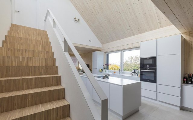 Ein moderner Küchen- und Treppenbereich mit hellem Holz und weißen Einbauten. Die Küche ist minimalistisch gestaltet, mit großen Fenstern, die natürliches Licht hereinlassen. 