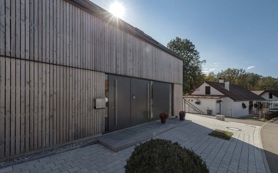 Modernes Gebäude mit vertikaler Holzfassade und großen Grautönen an Türen und Fenstern. Ein gepflasterter Zugang führt zur Haupteingangstür, neben der dekorative Pflanzgefäße stehen.