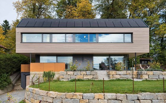 Ein rechteckiges, modernes Haus mit horizontaler Holzverkleidung und einer Photovoltaikanlage auf dem Dach. Das Gebäude hat eine erhöhte Lage mit einer Steinterrasse und ist von Bäumen umgeben