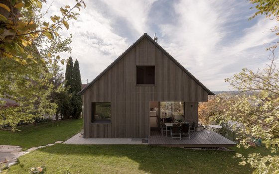 Ein schlichtes Giebelhaus mit vertikaler Holzverkleidung und einer kleinen Terrasse, die in den Garten führt. Die umliegenden Bäume und der grüne Rasen verstärken den ländlichen Charme des Hauses.