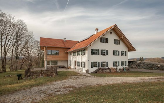 Szenisches Anwesen mit einer Kombination aus modernem und traditionellem Baustil, gelegen in einer idyllischen ländlichen Umgebung. Das Hauptgebäude ist weiß gestrichen mit charakteristischen Fensterläden, während der Anbau aus Naturholz besteht.