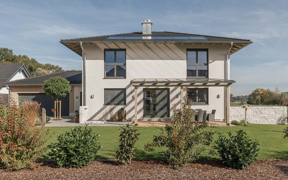 Zweistöckiges Einfamilienhaus mit hellgrauer Fassade, moderner Pergola über der Terrasse und gepflegtem Garten im Vordergrund. Große Fenster lassen viel Licht in das Haus.