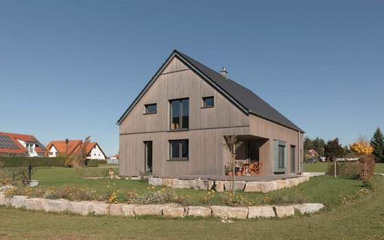 Ansicht eines modernen Wohnhauses mit grauer Holzverkleidung und spitzem Dach, gelegen in einer idyllischen Vorortsiedlung. Vorne im Bild sieht man eine schön angelegte Natursteinterrasse umgeben von einem blühenden Garten.
