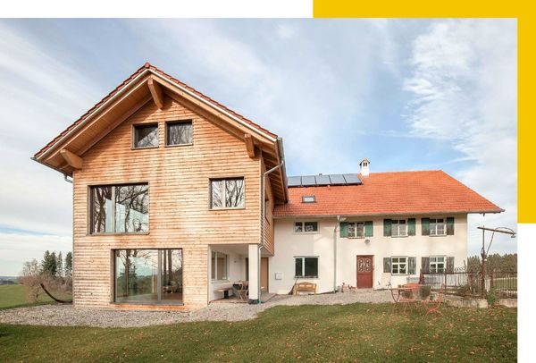 Ein modernes Bauernhaus mit rotem Satteldach, großem Fenster und Solarpaneelen. Der Garten ist weitläufig und von grüner Landschaft umgeben.