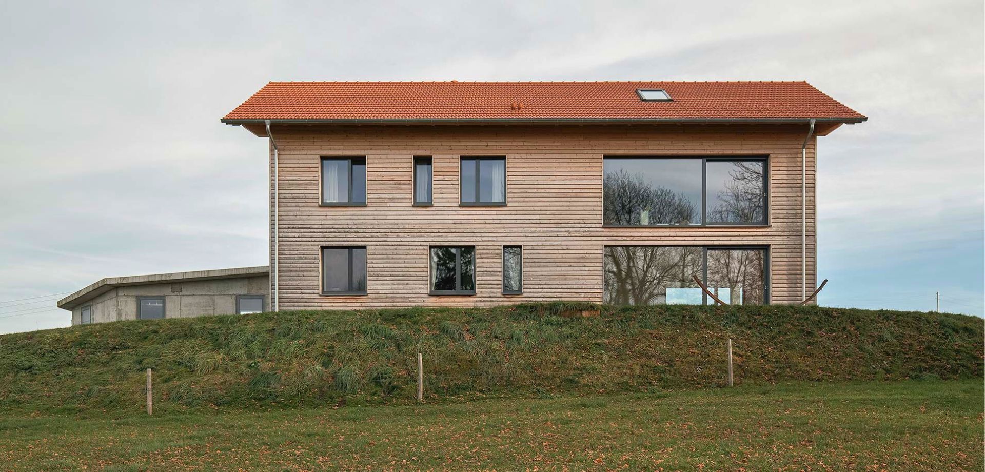 Ein zweistöckiges Haus mit Holzfassade und rotem Satteldach. Es steht auf einem grasbewachsenen Hügel und hat große Fenster, die den Innenraum erhellen.