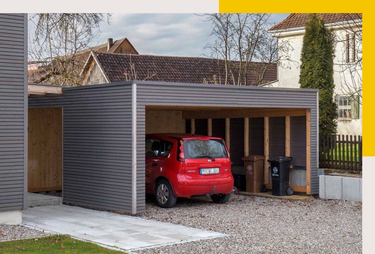 Rehklau-Referenz für Zimmerei-Arbeiten: Ein Carport mit grauer Holzverkleidung, der ein rotes Auto und mehrere Mülltonnen beherbergt.