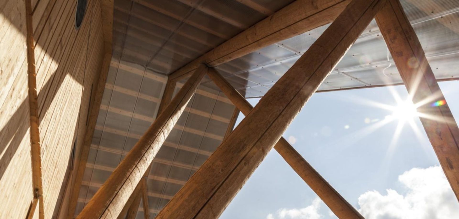 Nahaufnahme von Holzbalken, die eine Dachkonstruktion stützen. Sonnenstrahlen leuchten durch das Dach und erzeugen Lichtreflexe.