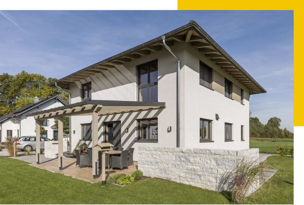 Rehklau-Referenz für Zimmerei-Arbeit: Modernes, weiß verputztes Haus mit Flachdach und großer Terrasse, auf der sich ein Tisch mit Stühlen befindet. Die Terrasse ist teils überdacht von einer Pergola, und das Haus steht auf einem gepflegten Rasen.