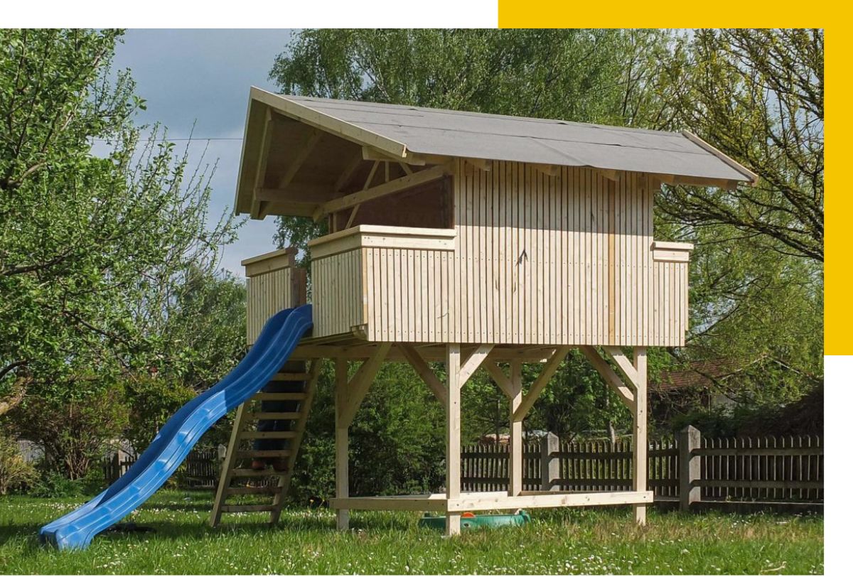 Rehklau Referenz für Zimmerei-Arbeit: Holzbaumhaus mit grauem Dach und blauer Rutsche im Garten, umgeben von grünen Bäumen und Wiese. Das Baumhaus hat einen offenen Bereich und ist über eine Holztreppe erreichbar.