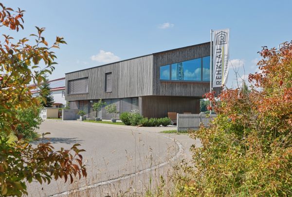 Ein rechteckiges Bürogebäude mit einer Fassade aus vertikalen Holzlatten. Ein großer blauer Fensterabschnitt reflektiert den Himmel, und im Vordergrund sind herbstliche Pflanzen sichtbar. Es stellt das Rehklau Vollholz Firmengebäude dar.