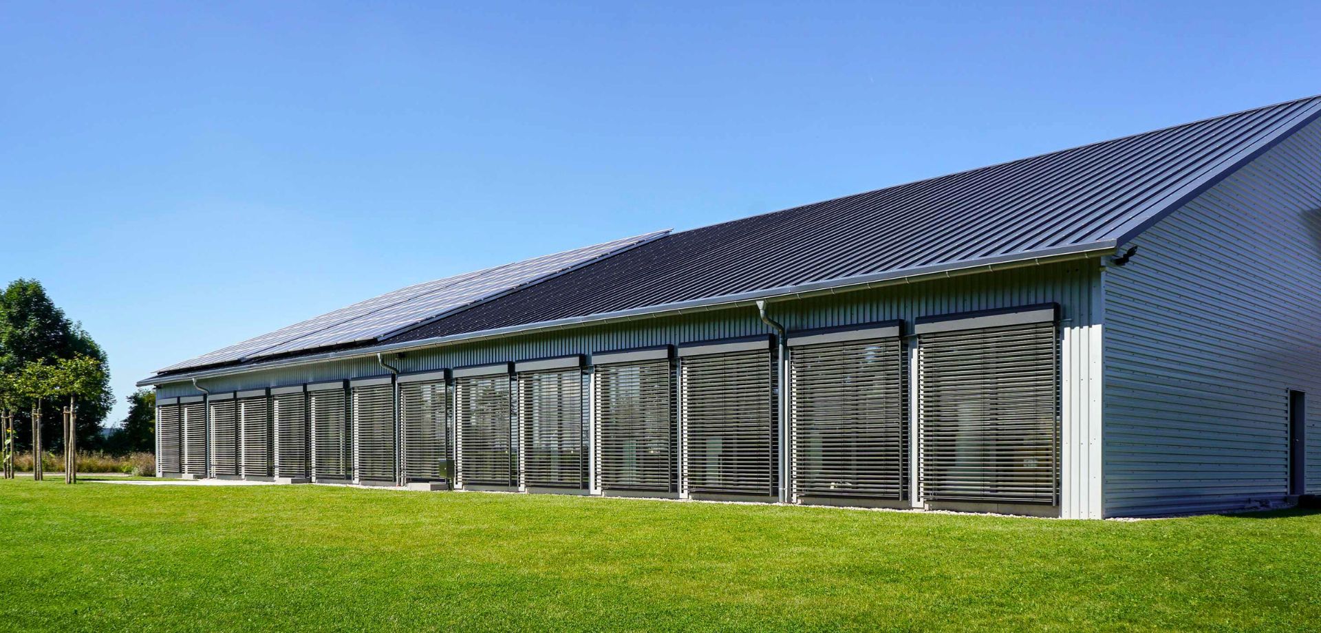 Ein langes, modernes Gebäude mit grauer Metallverkleidung und einer Reihe von großen Fenstern, die teilweise durch Rollos abgedunkelt sind. Das Gebäude steht auf einer grünen Wiese unter einem klaren blauen Himmel.